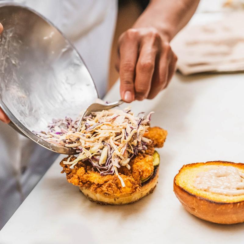 Hands topping a fried chicken sandwich with coleslaw using a fork