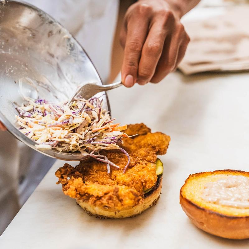 Hands adding coleslaw to a fried chicken sandwich with a fork