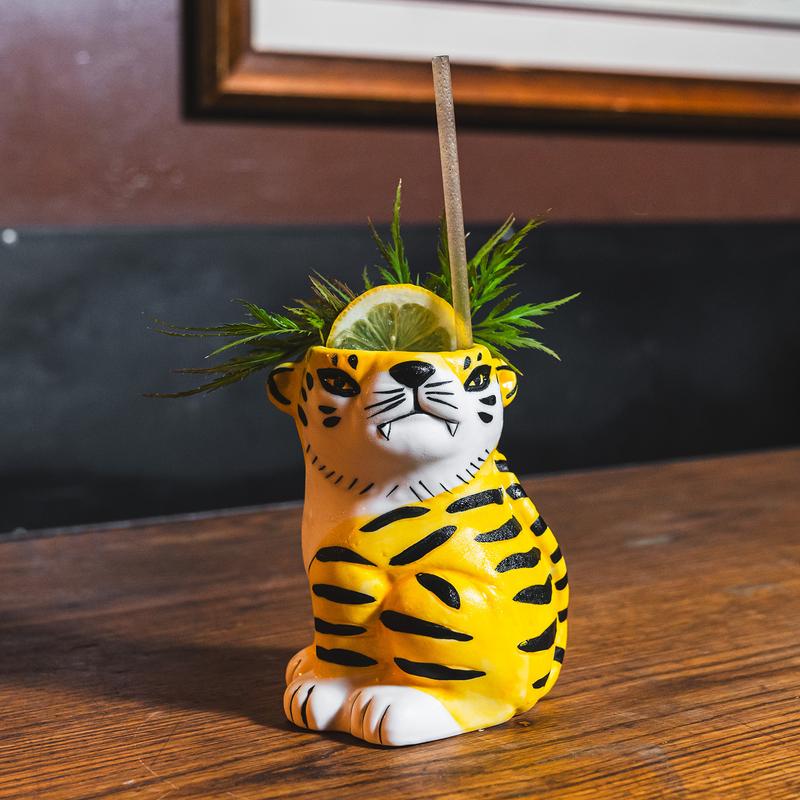 Cocktail in a tiger tiki mug garnished with a lemon slice and greenery