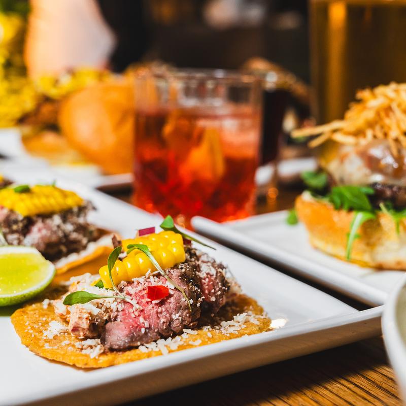 Carne asada tacos with a blurred cocktail and burgers on a table